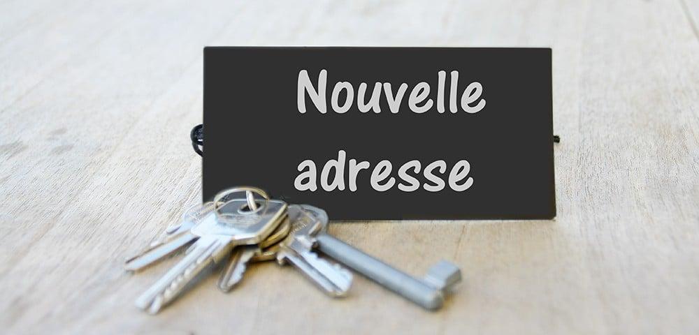 Keys resting on a wooden surface beside a small black sign that reads 'Nouvelle adresse' (French for 'New address').