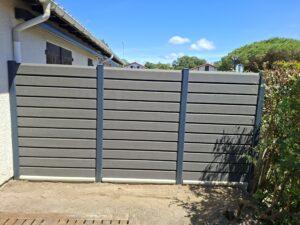 Gray horizontal-slat privacy fence along a dirt yard with a beige house and gutter on the left, blue sky above.