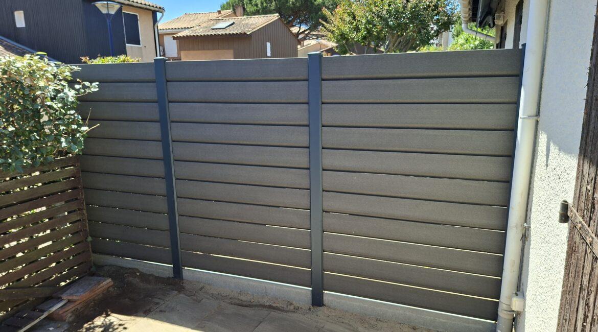 Grey horizontal-slatted fence enclosing a small backyard, with a blue sky, neighboring houses in the background, and a drainpipe along the right wall; plants and a wooden pallet are near the left side.