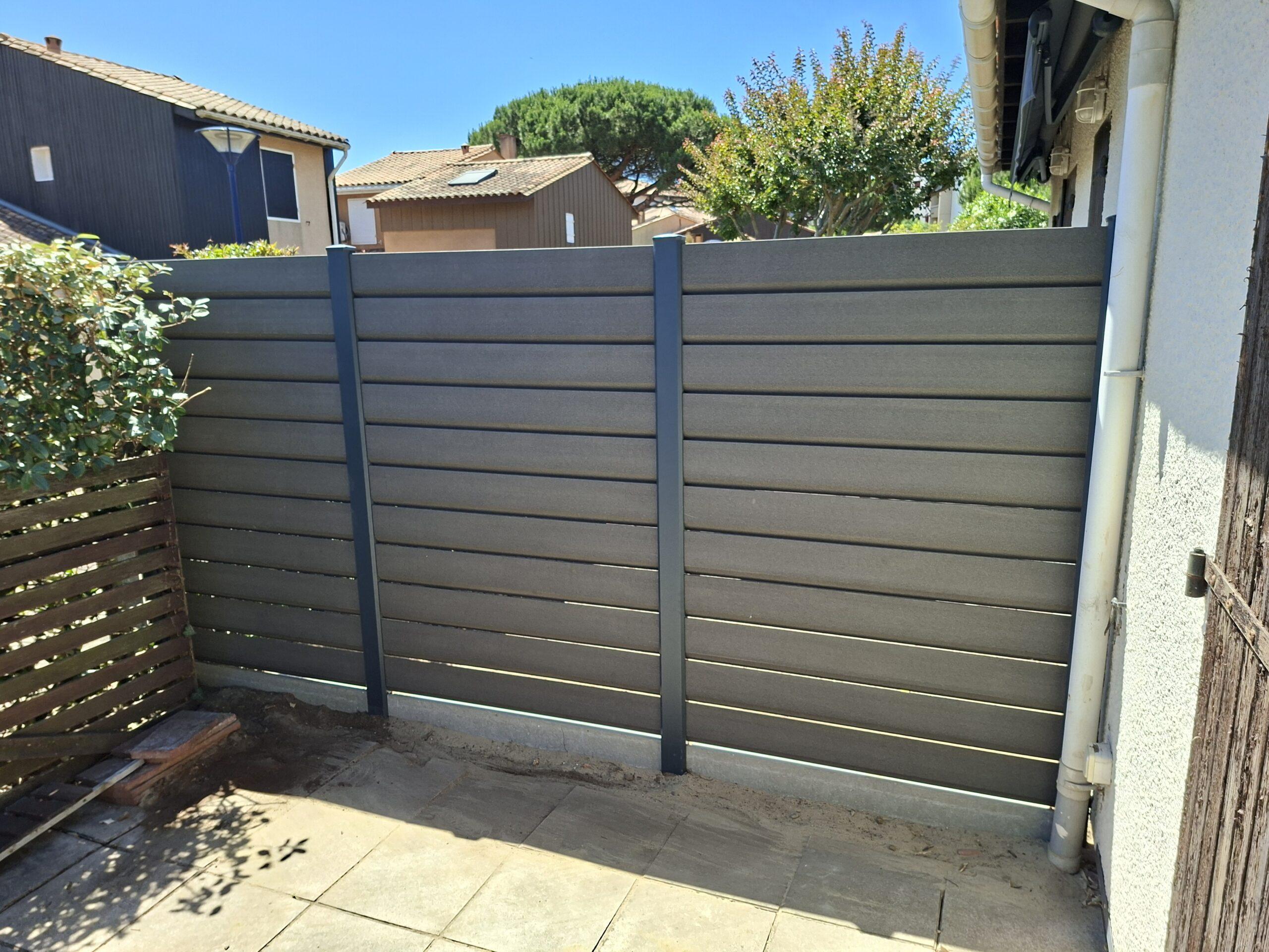 Grey horizontal-slatted fence enclosing a small backyard, with a blue sky, neighboring houses in the background, and a drainpipe along the right wall; plants and a wooden pallet are near the left side.