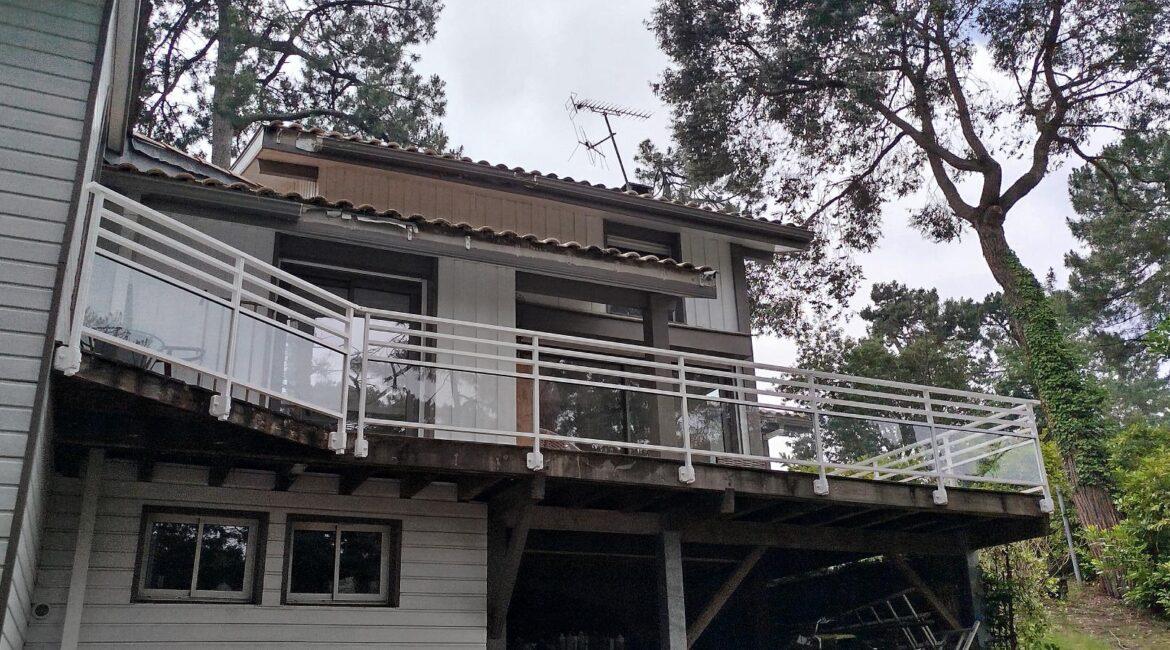 Two-story house with a glass-railed balcony extending from the second floor, surrounded by trees.