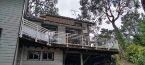 Two-story house with a glass-railed balcony extending from the second floor, surrounded by trees.
