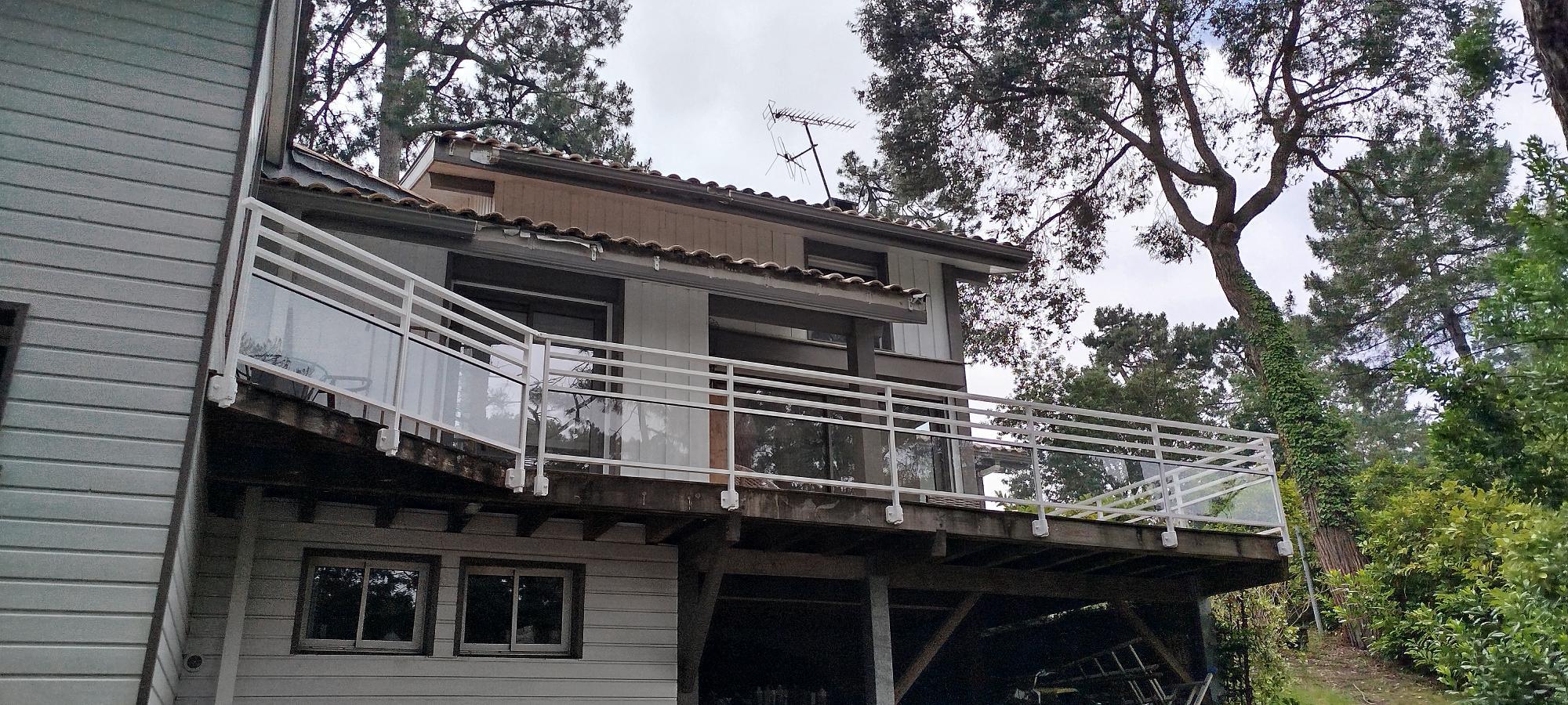 Two-story house with a glass-railed balcony extending from the second floor, surrounded by trees.
