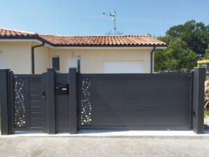 Dark gray decorative fence with circular perforations in front of a beige house with a terracotta-tiled roof and a roof antenna.