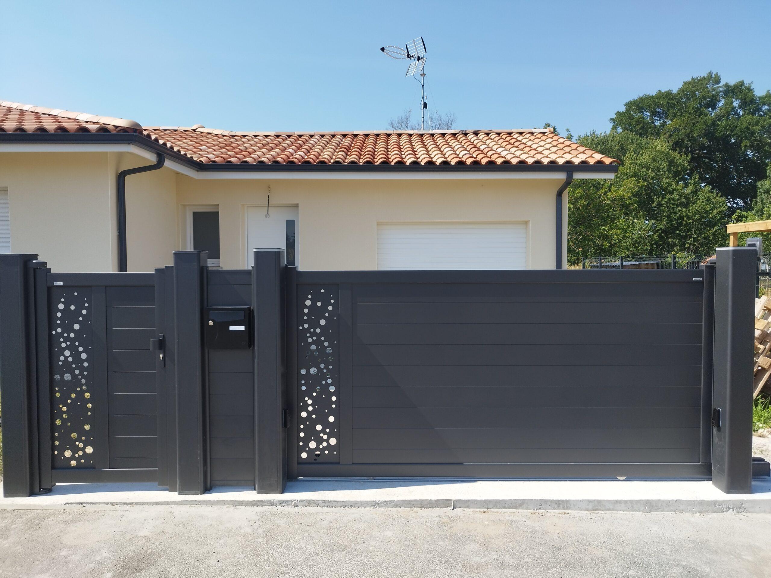 Dark gray decorative fence with circular perforations in front of a beige house with a terracotta-tiled roof and a roof antenna.