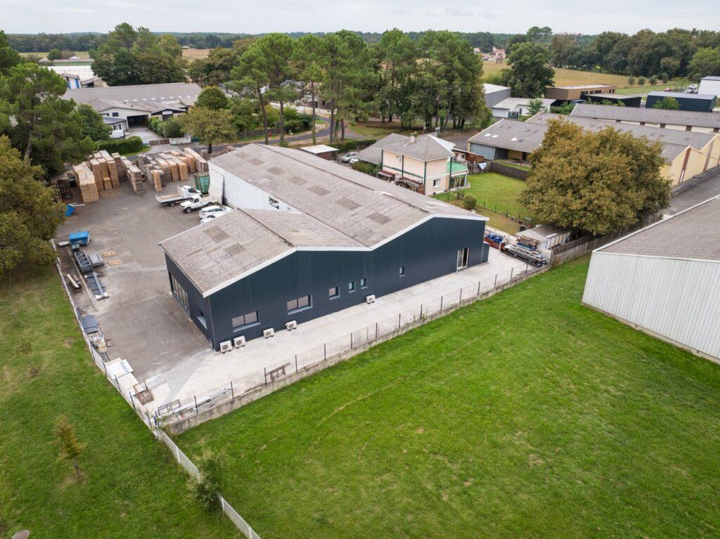 Aerial view of a warehouse complex with stacked pallets outside, loading trucks, and surrounding trees and fields.