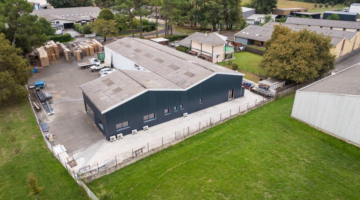 Aerial view of a warehouse complex with stacked pallets outside, loading trucks, and surrounding trees and fields.